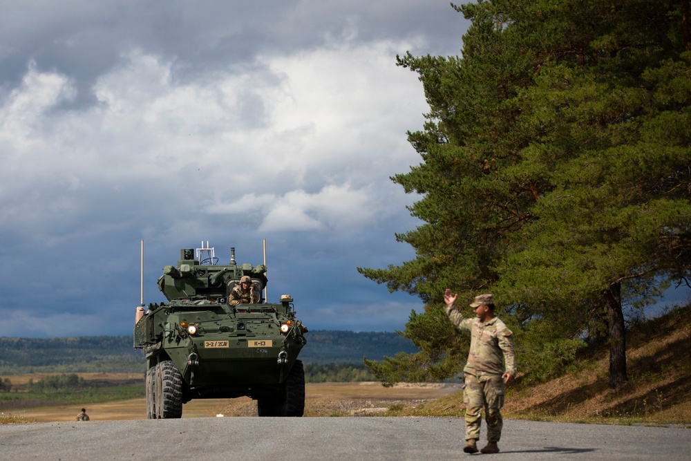 A U.S. Army Stryker armored fighting vehicle marked '3-2/2CW' and 'K-13' is positioned on a paved road in what appears to be a Nordic or Northern European training environment. A soldier in MultiCam/OCP camouflage uniform is directing the vehicle with a raised hand gesture. The scene appears to be a military exercise or movement operation.