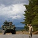 A U.S. Army Stryker armored fighting vehicle marked '3-2/2CW' and 'K-13' is positioned on a paved road in what appears to be a Nordic or Northern European training environment. A soldier in MultiCam/OCP camouflage uniform is directing the vehicle with a raised hand gesture. The scene appears to be a military exercise or movement operation.