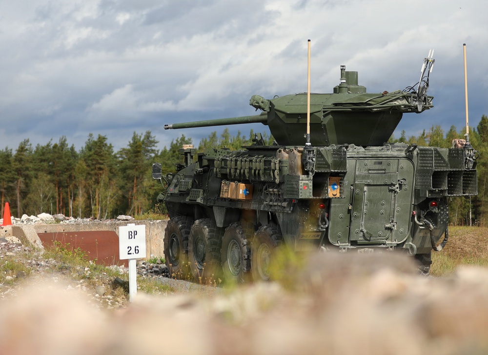 The image shows a wheeled armored fighting vehicle, consistent with a Stryker Mobile Gun System (MGS) or similar 8-wheeled armored vehicle, equipped with a large-caliber cannon turret and slat/cage armor on its sides. The vehicle is positioned on a training range or exercise area, indicated by the 'BP 2.6' marker post visible to the left. The setting appears to be a Northern European or Scandinavian environment based on the conifer tree line and terrain.