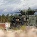 The image shows a wheeled armored fighting vehicle, consistent with a Stryker Mobile Gun System (MGS) or similar 8-wheeled armored vehicle, equipped with a large-caliber cannon turret and slat/cage armor on its sides. The vehicle is positioned on a training range or exercise area, indicated by the 'BP 2.6' marker post visible to the left. The setting appears to be a Northern European or Scandinavian environment based on the conifer tree line and terrain.