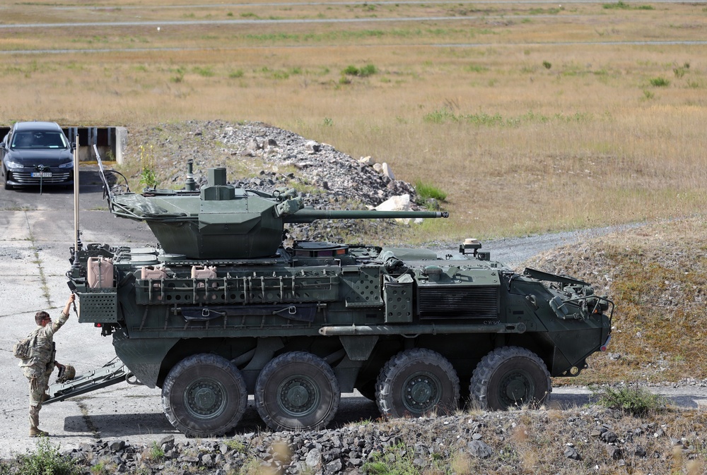 The image shows a large green 8-wheeled armored fighting vehicle, consistent with a Stryker-type infantry carrier variant, positioned on a paved surface in an open terrain area. A soldier in camouflage uniform is standing beside the vehicle, appearing to interact with equipment on its left side. A civilian car and a rubble berm are visible in the background.