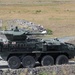 The image shows a large green 8-wheeled armored fighting vehicle, consistent with a Stryker-type infantry carrier variant, positioned on a paved surface in an open terrain area. A soldier in camouflage uniform is standing beside the vehicle, appearing to interact with equipment on its left side. A civilian car and a rubble berm are visible in the background.