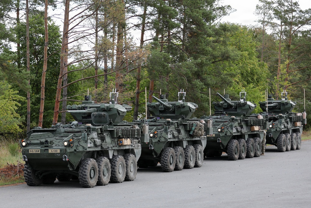 A column of four U.S. Army Stryker wheeled armored fighting vehicles is parked in formation on a paved road surrounded by a mixed forest. The vehicles appear to be equipped with Remote Weapon Stations (RWS) or turret-mounted weapon systems, consistent with the Stryker Infantry Carrier Vehicle (ICV) or Dragoon variant. Unit markings are visible on the lead vehicle, indicating affiliation with 3-2 Cavalry Regiment or similar unit.