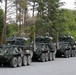 A column of four U.S. Army Stryker wheeled armored fighting vehicles is parked in formation on a paved road surrounded by a mixed forest. The vehicles appear to be equipped with Remote Weapon Stations (RWS) or turret-mounted weapon systems, consistent with the Stryker Infantry Carrier Vehicle (ICV) or Dragoon variant. Unit markings are visible on the lead vehicle, indicating affiliation with 3-2 Cavalry Regiment or similar unit.