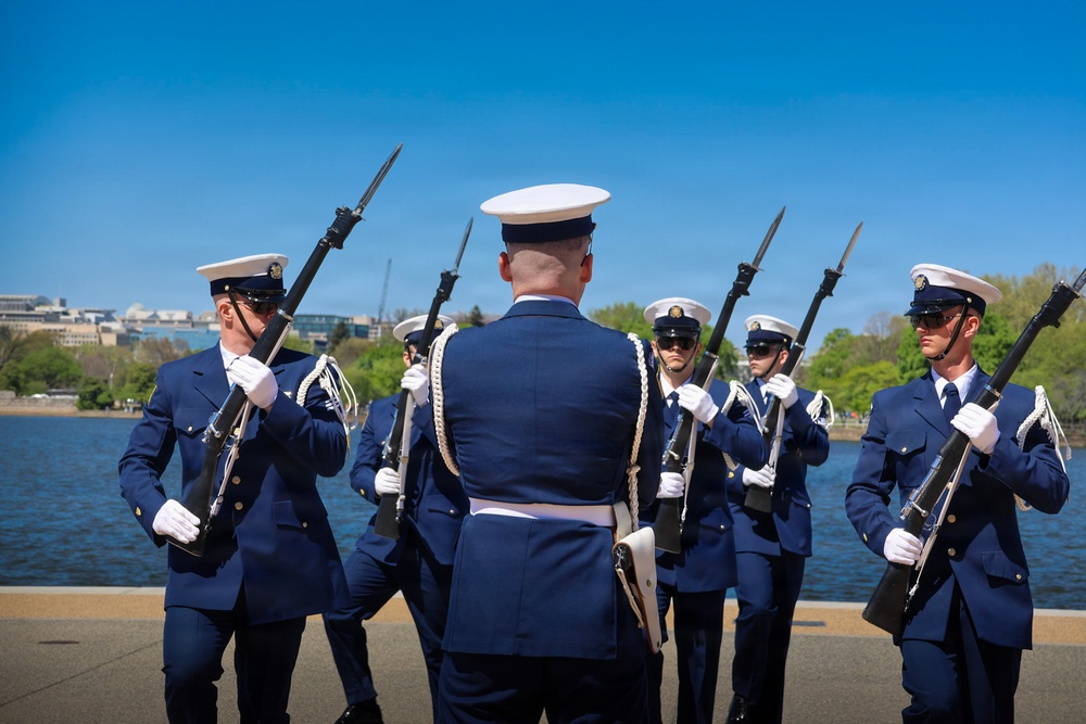 A military honor guard unit dressed in navy blue dress uniforms with white covers (caps) performs a ceremonial rifle drill near a waterfront, likely in Washington D.C. based on the recognizable Tidal Basin or Potomac River backdrop with city buildings visible. The personnel are carrying bayonet-fitted rifles and wearing white gloves, consistent with a formal ceremonial performance.