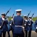 A military honor guard unit dressed in navy blue dress uniforms with white covers (caps) performs a ceremonial rifle drill near a waterfront, likely in Washington D.C. based on the recognizable Tidal Basin or Potomac River backdrop with city buildings visible. The personnel are carrying bayonet-fitted rifles and wearing white gloves, consistent with a formal ceremonial performance.