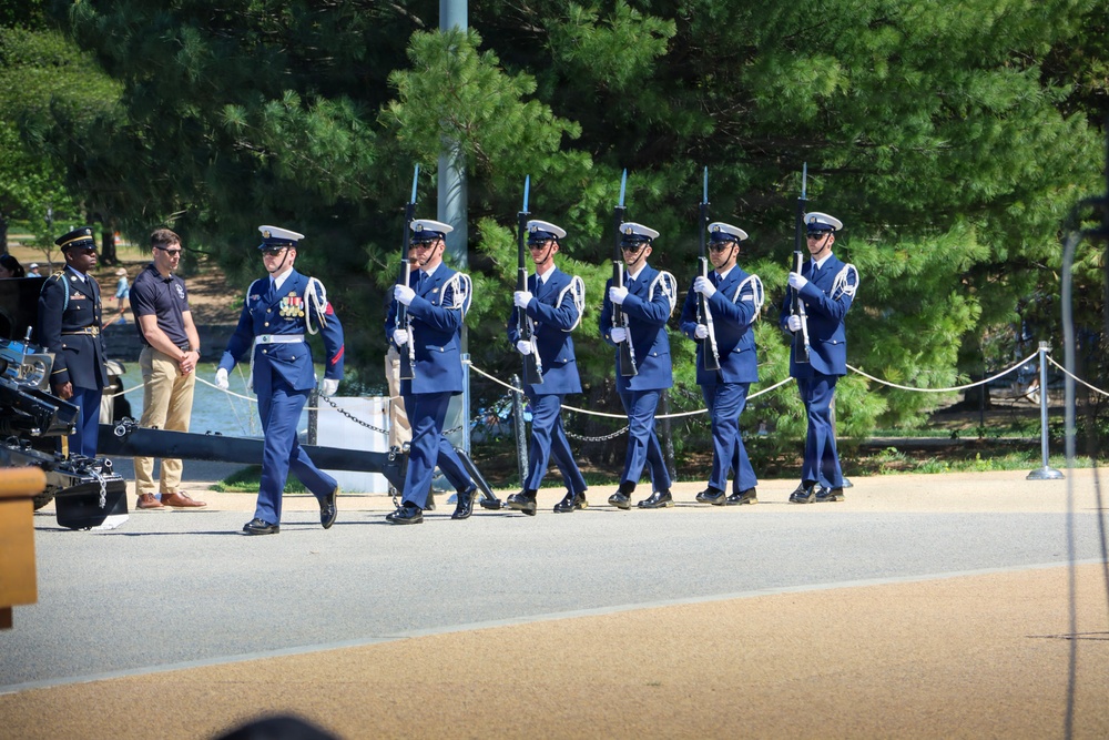 A military honor guard unit in dress blue uniforms is marching in formation while carrying rifles with bayonets attached. The ceremony appears to be taking place outdoors on a paved path, likely at a memorial or official venue, with spectators and other military personnel visible nearby. The soldiers are wearing white garrison caps and white gloves, consistent with a formal ceremonial drill.