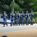 A military honor guard unit in dress blue uniforms is marching in formation while carrying rifles with bayonets attached. The ceremony appears to be taking place outdoors on a paved path, likely at a memorial or official venue, with spectators and other military personnel visible nearby. The soldiers are wearing white garrison caps and white gloves, consistent with a formal ceremonial drill.