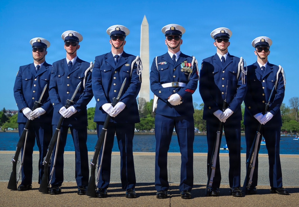 Six uniformed military personnel pose in formal dress uniforms in front of the Washington Monument and the Tidal Basin in Washington, D.C. Five members hold rifles with fixed bayonets at rest position, while the central figure — appearing to be the commanding officer — stands at ease wearing multiple medals and a white belt. The image appears to be an official or ceremonial photograph of a U.S. Coast Guard Honor Guard unit.