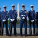 Six uniformed military personnel pose in formal dress uniforms in front of the Washington Monument and the Tidal Basin in Washington, D.C. Five members hold rifles with fixed bayonets at rest position, while the central figure — appearing to be the commanding officer — stands at ease wearing multiple medals and a white belt. The image appears to be an official or ceremonial photograph of a U.S. Coast Guard Honor Guard unit.