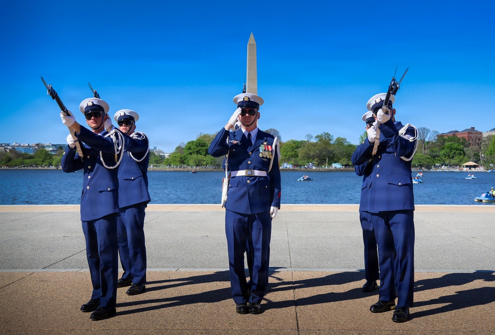 Five uniformed personnel, identifiable as U.S. Coast Guard members based on their distinctive blue dress uniforms and white covers (caps), are performing a ceremonial drill near the Tidal Basin in Washington, D.C. Four members are raising rifles with bayonets while one central figure renders a salute, suggesting a formal honor guard or ceremonial event. The Washington Monument is clearly visible in the background, confirming the location.