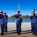 Five uniformed personnel, identifiable as U.S. Coast Guard members based on their distinctive blue dress uniforms and white covers (caps), are performing a ceremonial drill near the Tidal Basin in Washington, D.C. Four members are raising rifles with bayonets while one central figure renders a salute, suggesting a formal honor guard or ceremonial event. The Washington Monument is clearly visible in the background, confirming the location.