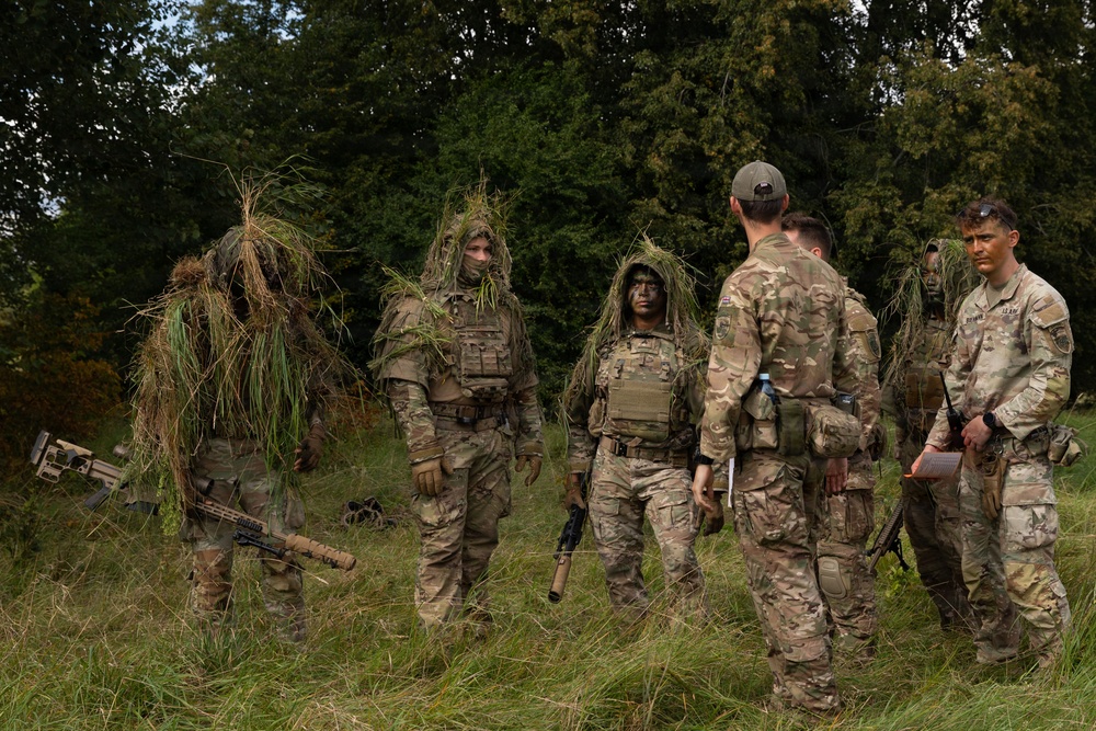 A group of military personnel in multicam/camouflage uniforms are gathered in an open grassy area near a tree line. Several soldiers are wearing ghillie suit-style camouflage coverings over their helmets and upper bodies, suggesting sniper or reconnaissance training. The group appears to be in a debrief or instruction session, with some personnel holding weapons and one individual holding what appears to be a notepad.