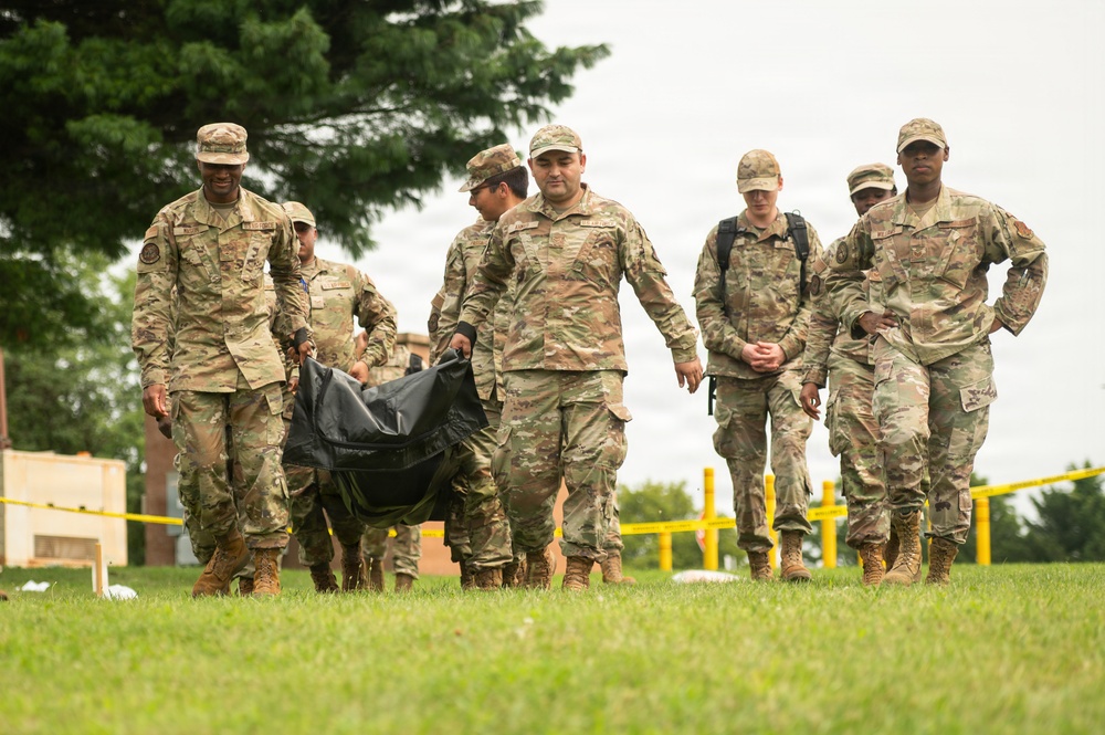A group of military personnel in MultiCam/OCP camouflage uniforms are walking across a grassy field during what appears to be a training exercise or field activity. Two soldiers in the foreground are carrying a black duffel bag or equipment bag between them. Yellow caution tape and bollards are visible in the background, suggesting a designated training or exercise area.