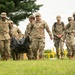 A group of military personnel in MultiCam/OCP camouflage uniforms are walking across a grassy field during what appears to be a training exercise or field activity. Two soldiers in the foreground are carrying a black duffel bag or equipment bag between them. Yellow caution tape and bollards are visible in the background, suggesting a designated training or exercise area.