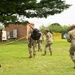 A group of military personnel in OCP (Operational Camouflage Pattern) uniforms are moving across a grassy area near a brick building. One soldier is carrying a black backpack while others appear to be walking toward a cordoned-off area marked with yellow caution tape. The setting appears to be a military installation or base during a training exercise or operational activity.