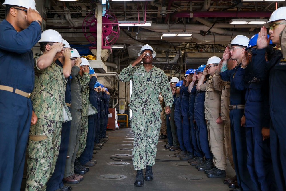 A U.S. Navy sailor in NWU Type I (digital camouflage) uniform and white hard hat walks through a ceremonial sideboy formation aboard what appears to be a naval vessel or shipyard facility. The central figure is returning a salute while two rows of personnel — in various uniforms and hard hats — render honors on both sides. This appears to be a 'walking the plank' or retirement/transfer ceremony, a traditional naval honors ritual.