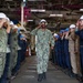 A U.S. Navy sailor in NWU Type I (digital camouflage) uniform and white hard hat walks through a ceremonial sideboy formation aboard what appears to be a naval vessel or shipyard facility. The central figure is returning a salute while two rows of personnel — in various uniforms and hard hats — render honors on both sides. This appears to be a 'walking the plank' or retirement/transfer ceremony, a traditional naval honors ritual.