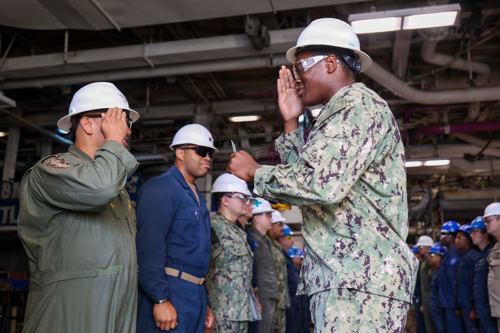 A military ceremony or inspection is taking place inside what appears to be a naval vessel's hangar bay or engineering space. Two personnel in the foreground are exchanging salutes, with one wearing Navy Working Uniform (NWU) digital camouflage and a white hard hat, and the other in an olive green flight suit. Multiple sailors and personnel in various uniforms and hard hats are lined up in the background.