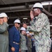 A military ceremony or inspection is taking place inside what appears to be a naval vessel's hangar bay or engineering space. Two personnel in the foreground are exchanging salutes, with one wearing Navy Working Uniform (NWU) digital camouflage and a white hard hat, and the other in an olive green flight suit. Multiple sailors and personnel in various uniforms and hard hats are lined up in the background.
