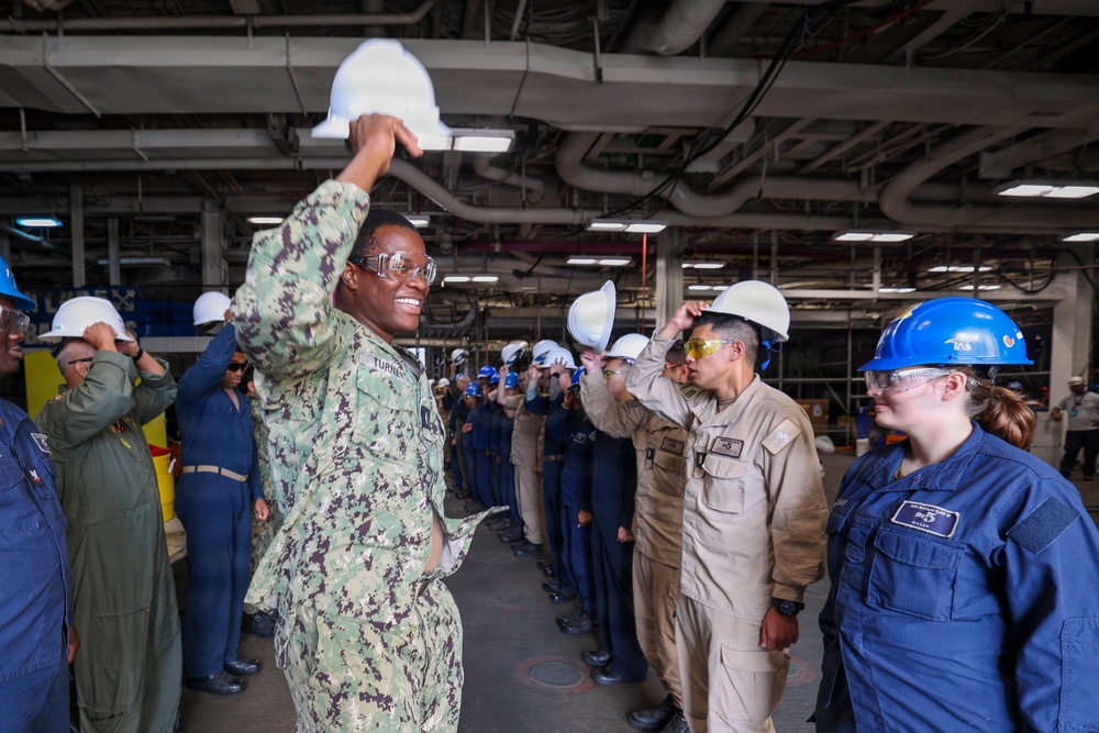 A group of military personnel and civilians are gathered in what appears to be the hangar bay or interior deck of a naval vessel. A central figure in U.S. Navy digital camouflage (NWU) uniform, identified by nametape as 'TURNER,' is smiling and raising a white hard hat above their head. The group appears to be participating in a safety equipment demonstration or orientation event.