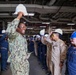 A group of military personnel and civilians are gathered in what appears to be the hangar bay or interior deck of a naval vessel. A central figure in U.S. Navy digital camouflage (NWU) uniform, identified by nametape as 'TURNER,' is smiling and raising a white hard hat above their head. The group appears to be participating in a safety equipment demonstration or orientation event.