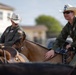 Two individuals wearing military flight suits (olive/dark green) and white cowboy hats are riding horses in what appears to be an outdoor setting. Both wear patches consistent with U.S. Air Force insignia, including an American flag patch visible on the left rider's sleeve. The combination of military uniforms and western cowboy attire suggests a themed event or military rodeo activity.