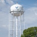 A herd of Texas Longhorn cattle walks along a paved road in front of a U.S. Air Force water tower. The tower is white with the U.S. Air Force logo and text prominently displayed. The scene juxtaposes traditional Western ranching culture with a modern military installation.