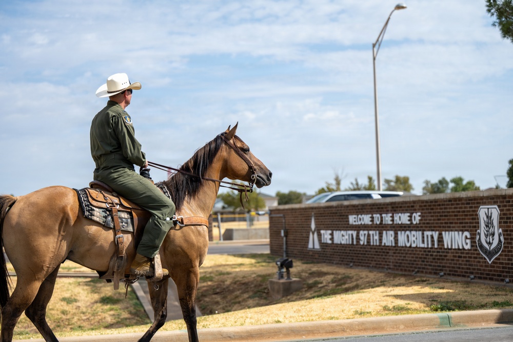 A person wearing a green military flight suit and white cowboy hat rides a buckskin horse in front of the entrance sign for the 97th Air Mobility Wing military installation. The brick wall sign reads 'Welcome to the Home of The Mighty 97th Air Mobility Wing' and displays the unit's insignia. The scene blends Western/cowboy culture with U.S. Air Force military identity.