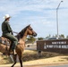 A person wearing a green military flight suit and white cowboy hat rides a buckskin horse in front of the entrance sign for the 97th Air Mobility Wing military installation. The brick wall sign reads 'Welcome to the Home of The Mighty 97th Air Mobility Wing' and displays the unit's insignia. The scene blends Western/cowboy culture with U.S. Air Force military identity.