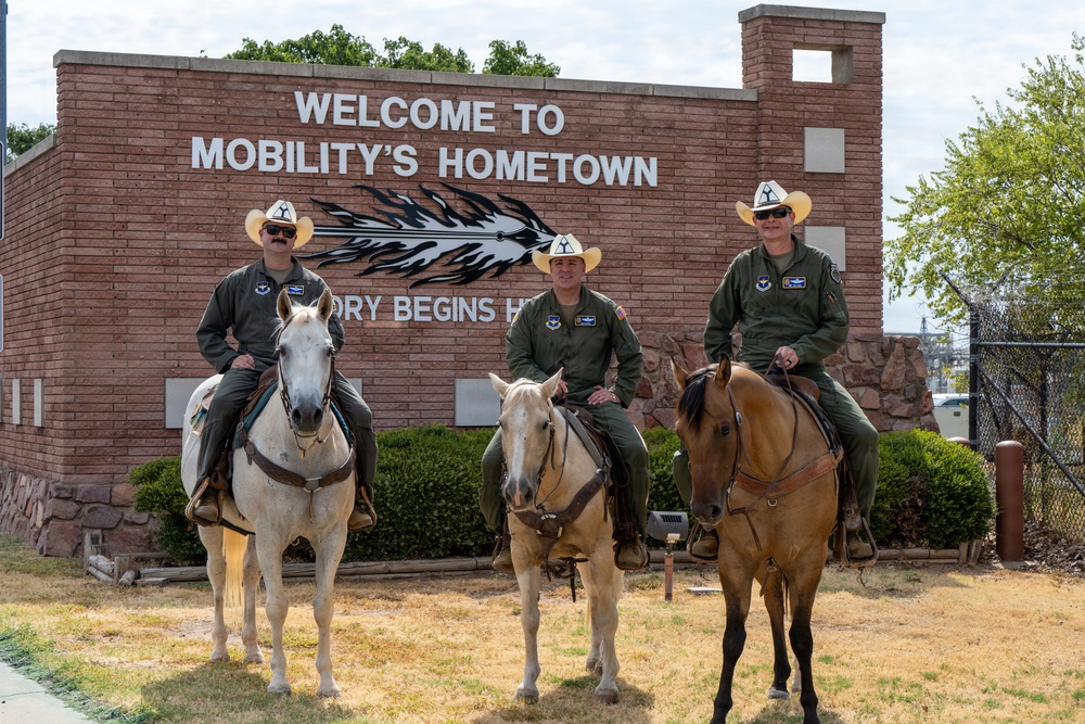 Three individuals wearing military flight suits (olive green) and white cowboy hats are posed on horseback in front of a brick sign reading 'Welcome to Mobility's Hometown' with partially visible text 'ory Begins H[ere]'. The riders appear to be U.S. Air Force personnel based on visible patches and insignia on their uniforms. The setting appears to be a military installation entrance, likely Scott Air Force Base in Illinois, known as 'Mobility's Hometown'.