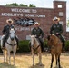 Three individuals wearing military flight suits (olive green) and white cowboy hats are posed on horseback in front of a brick sign reading 'Welcome to Mobility's Hometown' with partially visible text 'ory Begins H[ere]'. The riders appear to be U.S. Air Force personnel based on visible patches and insignia on their uniforms. The setting appears to be a military installation entrance, likely Scott Air Force Base in Illinois, known as 'Mobility's Hometown'.