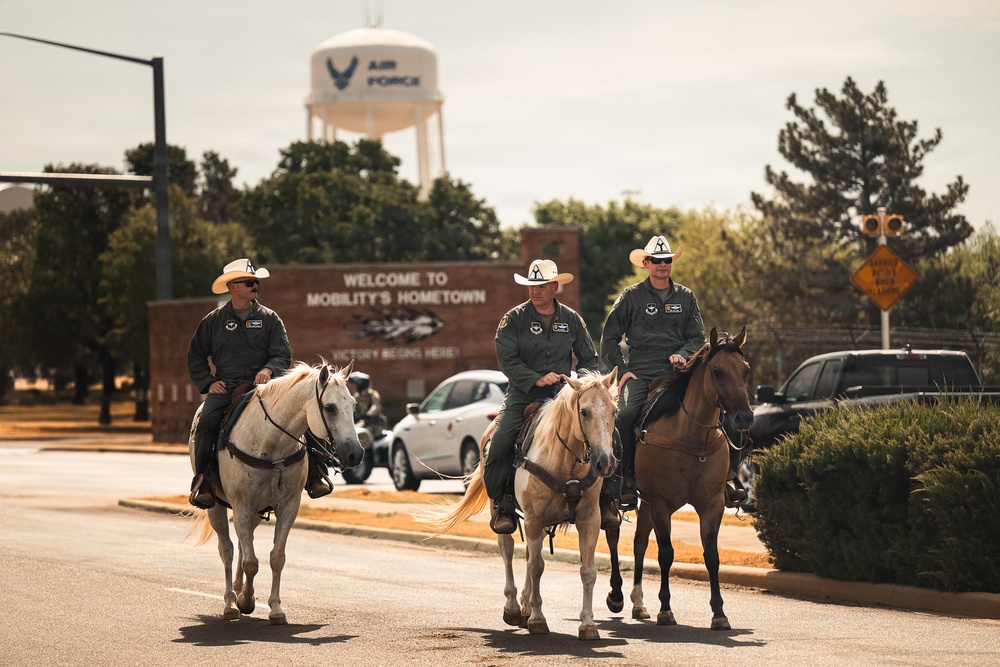 Three individuals wearing military flight suits and white cowboy hats ride horses down a road on what appears to be a U.S. Air Force base. A water tower prominently displaying 'Air Force' with the USAF symbol is visible in the background, along with a sign reading 'Welcome to Mobility's Hometown.' The scene blends military and Western cowboy aesthetics in what appears to be a ceremonial or celebratory event.