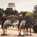 Three individuals wearing military flight suits and white cowboy hats ride horses down a road on what appears to be a U.S. Air Force base. A water tower prominently displaying 'Air Force' with the USAF symbol is visible in the background, along with a sign reading 'Welcome to Mobility's Hometown.' The scene blends military and Western cowboy aesthetics in what appears to be a ceremonial or celebratory event.