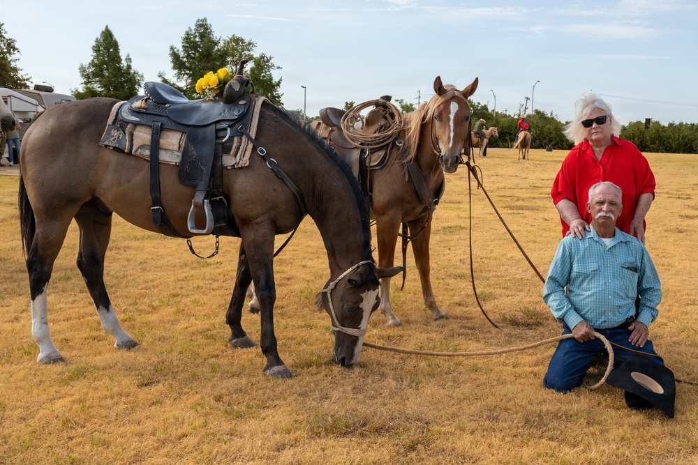 Two older adults pose with two saddled horses on a dry grass field. The man kneels in front holding a rope lasso and a cowboy hat, while the woman in a red shirt stands behind him with her hands on his shoulders. The setting appears to be an outdoor equestrian or rodeo event, with additional horses and riders visible in the background.