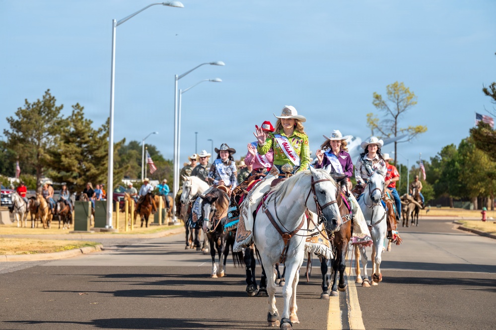 A western-style parade proceeds down a paved road featuring multiple riders on horseback, led by a young woman in a bright green shirt wearing a pageant sash and white cowboy hat riding a white horse. Several participants appear to be rodeo royalty contestants or queens, identifiable by their decorative sashes and ornate western attire. American flags are visible in the background along a tree-lined street with modern lamp posts.