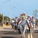 A western-style parade proceeds down a paved road featuring multiple riders on horseback, led by a young woman in a bright green shirt wearing a pageant sash and white cowboy hat riding a white horse. Several participants appear to be rodeo royalty contestants or queens, identifiable by their decorative sashes and ornate western attire. American flags are visible in the background along a tree-lined street with modern lamp posts.