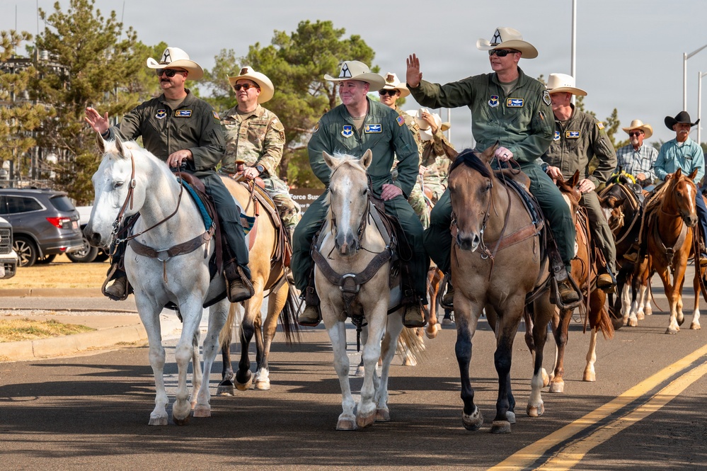 A group of military personnel wearing green flight suits and cowboy hats are riding horses along a paved road, appearing to participate in a ceremonial parade or procession. Several riders are waving to an unseen crowd, and military unit patches are visible on their uniforms. The scene blends military and Western/cowboy culture, likely at or near a military installation.