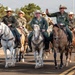 A group of military personnel wearing green flight suits and cowboy hats are riding horses along a paved road, appearing to participate in a ceremonial parade or procession. Several riders are waving to an unseen crowd, and military unit patches are visible on their uniforms. The scene blends military and Western/cowboy culture, likely at or near a military installation.