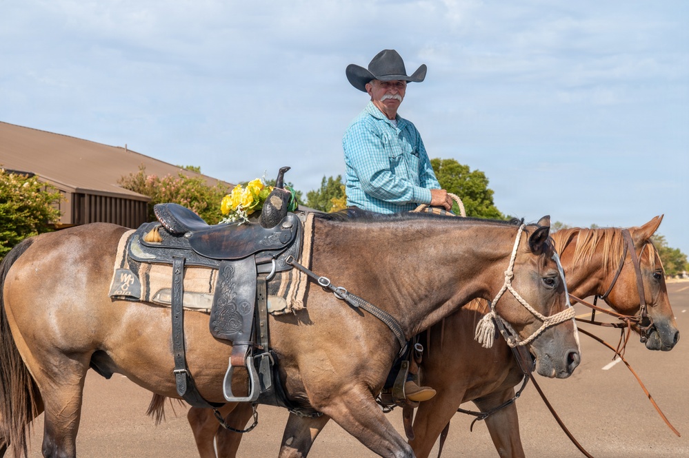 An older man wearing a black cowboy hat and turquoise plaid shirt rides a brown horse while leading a second saddled horse alongside him. The riderless horse carries a decorative western saddle adorned with yellow flowers, suggesting a ceremonial or memorial context such as a parade or tribute ride. The scene takes place on a paved road with trees and a building visible in the background.
