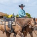 An older man wearing a black cowboy hat and turquoise plaid shirt rides a brown horse while leading a second saddled horse alongside him. The riderless horse carries a decorative western saddle adorned with yellow flowers, suggesting a ceremonial or memorial context such as a parade or tribute ride. The scene takes place on a paved road with trees and a building visible in the background.