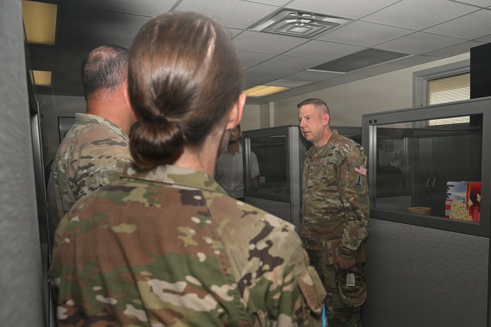 Three military personnel in OCP (Operational Camouflage Pattern) uniforms are gathered in what appears to be an office or administrative area with cubicle partitions. The soldier facing the camera appears to be speaking or briefing the other two individuals. The setting suggests an indoor military facility or headquarters environment.