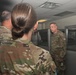 Three military personnel in OCP (Operational Camouflage Pattern) uniforms are gathered in what appears to be an office or administrative area with cubicle partitions. The soldier facing the camera appears to be speaking or briefing the other two individuals. The setting suggests an indoor military facility or headquarters environment.