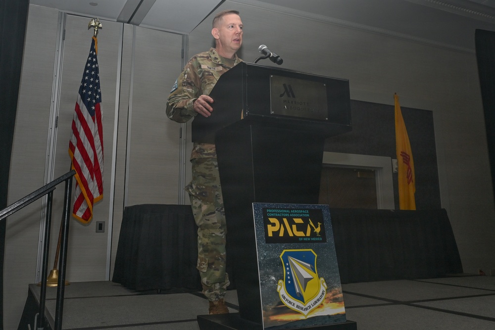 A military officer in camouflage uniform is delivering a speech at a podium inside what appears to be a Marriott hotel ballroom. The event is associated with the Professional Aerospace Contractors Association (PACA) of New Mexico and the Air Force Research Laboratory. Two flags are visible in the background — the United States flag and what appears to be the New Mexico state flag.