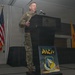 A military officer in camouflage uniform is delivering a speech at a podium inside what appears to be a Marriott hotel ballroom. The event is associated with the Professional Aerospace Contractors Association (PACA) of New Mexico and the Air Force Research Laboratory. Two flags are visible in the background — the United States flag and what appears to be the New Mexico state flag.