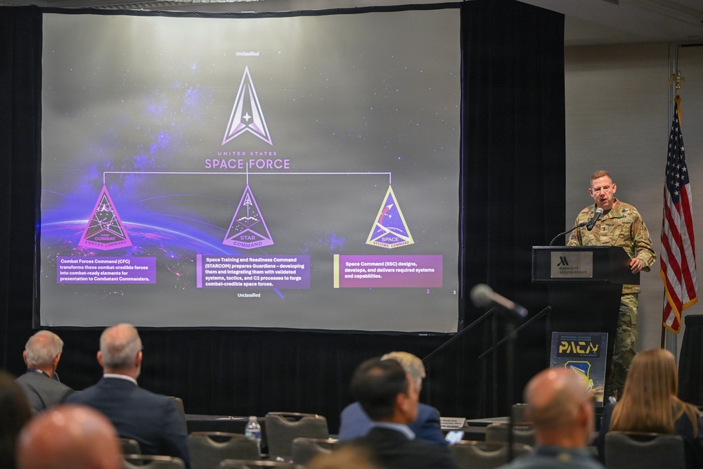 A military officer in combat uniform is presenting a briefing about the United States Space Force organizational structure at what appears to be a Marriott conference venue. The presentation slide displays the Space Force logo and three subordinate commands: Combat Forces Command (CFC), Space Training and Readiness Command (STARCOM), and Space Systems Command (SSC). An audience of civilians and officials is seated facing the presenter.
