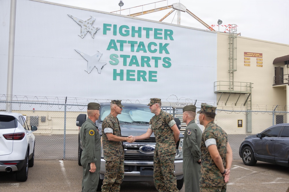 Military personnel in U.S. Marine Corps MARPAT camouflage uniforms and olive drab flight suits are gathered in a parking lot in front of a large building displaying a banner reading 'FIGHTER ATTACK STARTS HERE' with jet aircraft silhouettes. Two individuals are visibly shaking hands, suggesting a formal greeting or official visit. The setting appears to be a military aviation facility, likely a Marine Corps air station.
