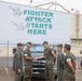Military personnel in U.S. Marine Corps MARPAT camouflage uniforms and olive drab flight suits are gathered in a parking lot in front of a large building displaying a banner reading 'FIGHTER ATTACK STARTS HERE' with jet aircraft silhouettes. Two individuals are visibly shaking hands, suggesting a formal greeting or official visit. The setting appears to be a military aviation facility, likely a Marine Corps air station.