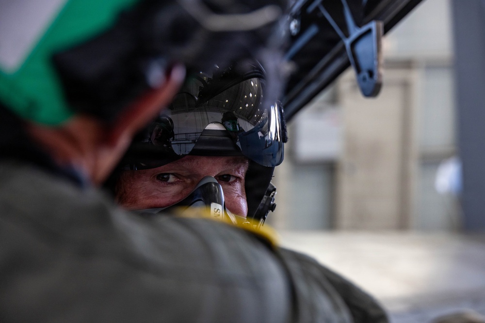 A close-up photograph of a military pilot or aircrew member wearing a flight helmet with a raised dark visor and an oxygen mask. The image is taken from behind a second person wearing a green helmet, creating a shallow depth-of-field effect. The setting appears to be inside or near a hangar or aircraft facility.
