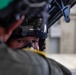 A close-up photograph of a military pilot or aircrew member wearing a flight helmet with a raised dark visor and an oxygen mask. The image is taken from behind a second person wearing a green helmet, creating a shallow depth-of-field effect. The setting appears to be inside or near a hangar or aircraft facility.